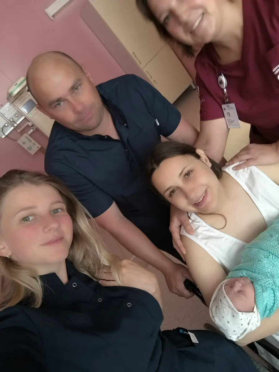 Селфи в палате: четверо взрослых вокруг женщины в белом, которая держит новорождённого в зелёном пледе